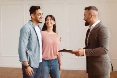 Gülümseyen Profesyonel Takım Elbiseli Emlakçının Portresi Genç Arap Çifte Yeni Daire Gösteriyor, Ellerinde Pano Tutuyorlar. Mutlu Orta Doğulu Adam ve Bayan Satıcıyla Kucaklaşıp Konuşuyor