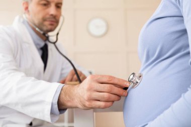 Bir doktor konseptini ziyaret et. Erkek jinekoloğun, hamile kızı steteskopla muayene edip kontrol ettiği portreyi kapat.