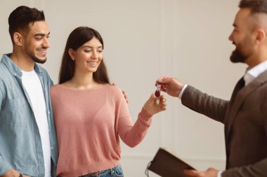 Buying New Apartment. Professional real estate agent wearing suit holding in hand and giving keys to young millennial Arab buyers. Happy woman taking key of future new apartment, selective focus