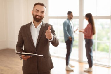 Başarılı bir anlaşma, Ev Sahipliği Konsepti. Gülümseyen profesyonel emlakçının portresi Başparmağı işaret gibi gösteren, seçici odaklanma. Bulanık Arkaplanda Konuşan Alıcılar Çifti