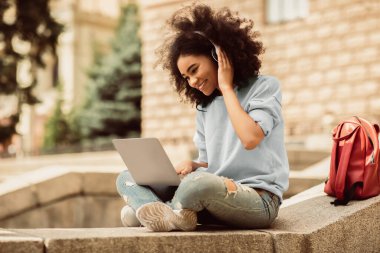 E-öğrenme. Diz üstü bilgisayar kullanan, kulaklık takan ve dışarıda oturup sanal eğitim dersi izleyen siyah bir kız öğrenci. Uzaktan Üniversite Eğitim Konsepti. Yan Görünüm