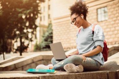 E-öğrenme. Dizüstü bilgisayarı olan siyah bir kız öğrencinin yan görüntüsü ya da dışarıda oturan kulaklık ve gözlük takan çevrimiçi bir konferans. Uzaklık Üniversite Eğitimi