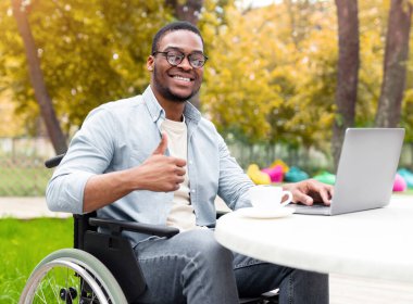 Engelliler için internet işi. Tekerlekli sandalyedeki gülümseyen özürlü adam dizüstü bilgisayarla çalışıyor, sonbahar parkındaki kafede baş parmak hareketi gösteriyor, uzaktan iş öneriyor, kopyalama alanı öneriyor.