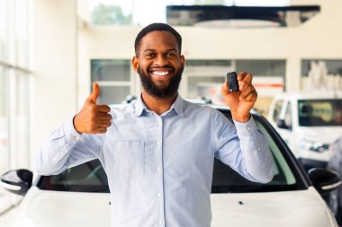 Neşeli Afrikalı Amerikalı adam yeni arabasının yanında duruyor, anahtarı tutuyor ve başparmağını kaldırıyor. Satış bürosunda yeni araç satın alımını kutlayan heyecanlı siyahi adam kameraya bakıp gülümsüyor.