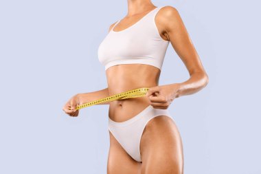 Sarı bantlı, beyaz iç çamaşırlı, yarı dönüşmüş, ince bir kadının yakın görüntüsü. Tanınmayan bir kadın mavi stüdyo duvarında izole edilmiş kilo kaybı diyeti ya da liposuction sonuçlarını kontrol ediyor.