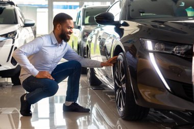 Gülümseyen Siyah Adam Salon 'da yeni araba alırken tekerlek ve lastikleri inceliyor, Afro-Amerikan erkek müşteri galeri merkezinde yeni araç seçiyor, Showroom' da çömelmiş vaziyette oturuyor.