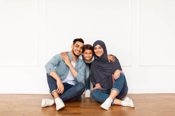 Portrait of loving happy Middle East family with little preschooler kid sitting near white wall looking at camera and smiling, parents enjoy time with small child posing together in modern new home