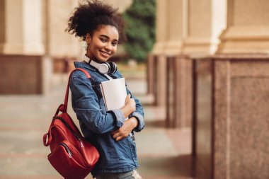 Üniversitenin kampüs bölgesinde sırt çantası ve kitaplarla poz veren başarılı siyahi kız kameraya gülümsüyor. Öğrencilik Yaşam Tarzı ve Modern Eğitim Konsepti