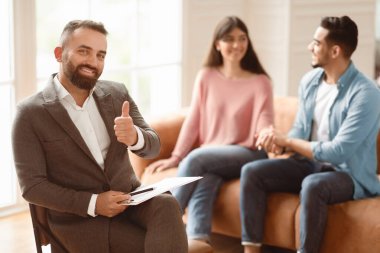 Ön plandaki profesyonel erkek psikolog, ofiste genç bir çiftle çalışan işaret dilini işaret ediyor. Eşler klinikte evlilik danışmanıyla görüşüyor.