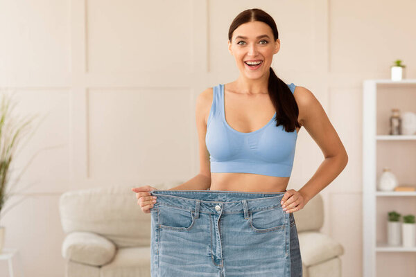 Тощая женщина по сравнению со старыми джинсами негабаритного размера после диеты и успешной потери веса стоять дома. Свободное пространство