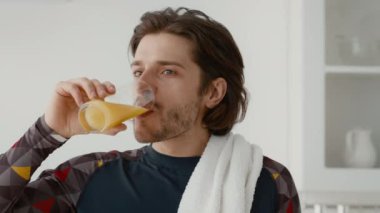 Genç beyaz bir adam ev çalışmasını tamamladıktan sonra sağlıklı bir portakal suyu yudumlayarak kendini tazeler. Boynunda bir havluyla odaklanmış ve gençleşmiş görünüyor..