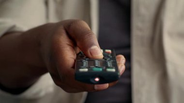 Genç bir Afrikalı Amerikalı adam evinde rahatlıyor, uzaktan kumandaya odaklanıyor, cihazlar ve modern teknolojiyle çevrili, tipik teknoloji kullanımı sergiliyor,