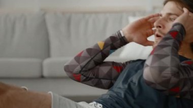 Genç beyaz bir adam evde egzersiz yapıyor, çekirdek egzersizleri yapıyor. Odaklanmış ve kendini adamış. İyi ışıklandırılmış bir alanda fitness 'e proaktif bir yaklaşım sergiliyor..