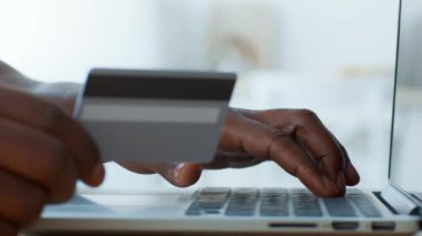 Genç bir Afro-Amerikan adam evinde oturuyor, dizüstü bilgisayarına odaklanmış. Bir elinde kredi kartı tutarken diğer elinde ise internet üzerinden alışveriş veya alışveriş yapıyor..