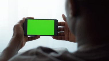 Afro-Amerikalı bir adam evde otururken elinde parlak yeşil ekranlı bir akıllı telefon tutuyor. Modern bir dijital yaşam tarzının altını çizerek cihazın içine çekilmiş gibi görünüyor..