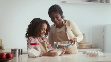 Bir anne ve kızı, sıcak mutfaklarında hamur işleri için malzemeleri karıştırarak mutlu bir şekilde birlikte çalışırlar. Annesi kâseye süt dökerken kız gülümsüyor ve neşeli bir yemek pişirme anı yaratıyor..