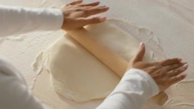 Afro-Amerikalı bir kadın mutfağında ahşap bir oklava ile hamuru dikkatlice yuvarlıyor. Konforlu mutfak iyi aydınlatılmış, pişirme ve yemek yapma tutkusunu sergiliyor..