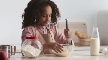 Sıcak bir mutfakta, genç bir kız hamur işi için malzemelerini hazırlarken fırın karışımını tatmaktan zevk alır.