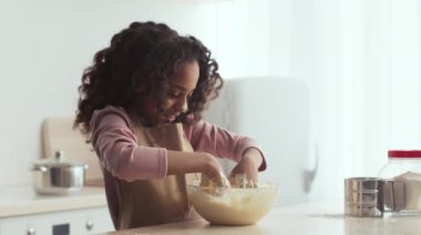 Parlak bir mutfakta, genç bir kız yemek pişirme süreciyle meşgul olan malzemeleri bir kasede karıştırmaktan hoşlanır.