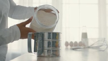 Bin yıllık Afro-Amerikan bir kadın kendi mutfağında pişirmeye odaklanmış durumda. O dikkatlice bir kavanozdan elek içine un ölçer, lezzetli bir ikram için malzemeleri hazırlar..