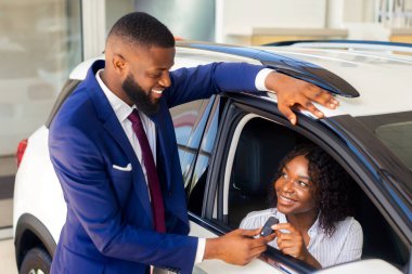 Mutlu siyahi kadın araba anahtarlarını ofis müdüründen alıyor keyifli Afrikalı Amerikalı kadın lüks bir aracın içinde oturuyor ve camdan gülümsüyor yeni otomobil satın almanın keyfini çıkartıyor,