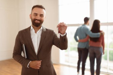 Neşeli Profesyonel Gayrimenkul Ajanı Pano Tutarken ve Anahtarları Elde Gösterirken Seçici Odaklanma Portresi. Yeni evliler yeni bir ev seçiyor. Pencereden dışarı bakıyor. Bulanık arka planda sarılıyorlar.