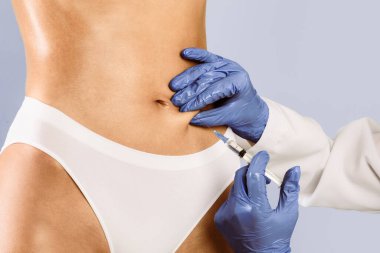 Koruyucu eldivenli plastik cerrah, göbek bölgesinde tanınmayan bir bayana iğne yapıyor. Mavi stüdyo geçmişi var. İnce bir kadın güzellik salonunda lipolizis tedavisi görüyor.