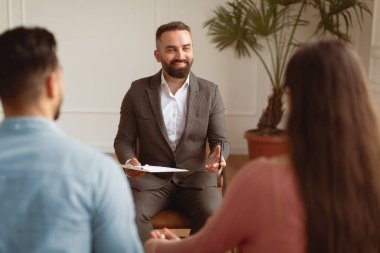 Smiling therapist talking with young millennial patients, back view of happy married couple spouses sitting on sofa holding hands visiting professional family counselor, resolving issues