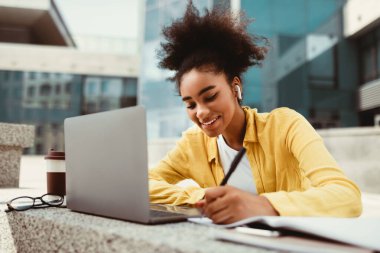 Gülümseyen siyah öğrenci dizüstü bilgisayarında not alıyor internetten öğreniyor ve ödevlerini yapıyor Modern Üniversite İnşaat Kampüsü 'nde oturuyor. Uzaklık Eğitimi, E-Öğrenme Konsepti