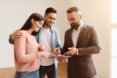 Gülümseyen Ev Danışmanının portresi modern daireleri genç Arap müşterilere gösteriyor, dijital tableti projeyle işaret ediyor, ev planını tartışıyor, Orta Doğulu mutlu bir aileye ev satışı yapıyor.
