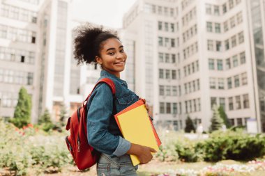 Başarılı bir öğrenci. Afro-Amerikan Kolej Kızı, elinde kitaplarla kameraya gülümseyen, dışarıda modern üniversite binasının yanında duran eğlenceli öğrencilere poz veriyor. Eğitim Konsepti