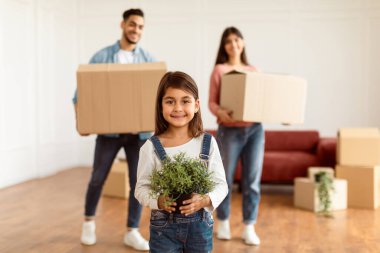 Ev Taşıma ve Taşınma Konsepti. Üç Evli Neşeli Aile Portresi, Mutlu Gülümseyen Küçük Kız Kucağına Odaklan, Bulanık Arkaplan 'da karton kutular taşıyan Ebeveynler