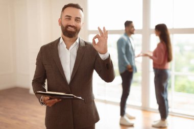 Başarılı bir anlaşma, Ev Sahipliği Konsepti. Gülümseyen Profesyonel Emlak Ajansı 'nın portresi İşaret İşareti, Seçici Odaklanma Gösteriyor. Bulanık Arkaplanda Konuşan Alıcılar