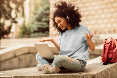 Uzaktan kumandalı sınıf binasının dışında oturan Afrikalı Amerikalı kız öğrenci dizüstü bilgisayar kullanarak video görüşmesi yapıyor. Uzaklık Eğitimi ve E-Öğrenme. Modern İletişim. Yan Görünüm