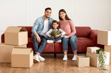 Ev Taşıma ve Yenileme Konsepti. Üç kişilik mutlu bir ailenin portresi. Yeni boş bir dairede oturmuş kameraya bakıyor ve poz veriyor.