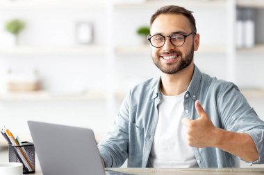 Masasında Türk bayrağıyla oturan mutlu genç adam dizüstü bilgisayar kullanıyor, evde el hareketi yapıyor. Çevrimiçi dış eğitim, göç ve vatandaşlık, yurtdışında çalışma kavramı