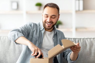 Memnun bir alıcı. Paketi alan mutlu beyaz adamın portresi, kutuları açmak, oyun kumandasını çıkarmak, oturma odasındaki kanepede oturmak. Milenyum çocuğu internetten satın almaktan memnun.