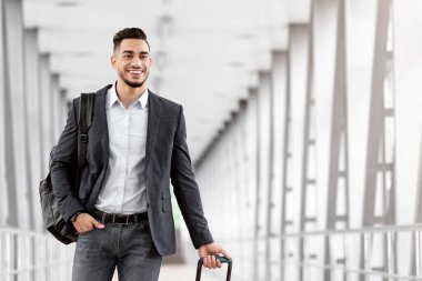Havaalanı terminalinde bavulla yürüyen genç bir orta doğunun portresi, biniş kapısına giderken bavul taşıyan gülümseyen Arap adam, hava yolculuklarının ve iş seyahatlerinin tadını çıkarıyor, kopyalama alanı.
