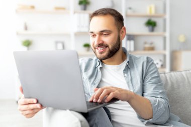 Neşeli bir milenyum adamı dizüstü bilgisayarla koltukta oturuyor. Ev ofisinden çalışıyor ya da internetten çalışıyor. Neşeli beyaz adam web konferansına katılıyor, video konferansına katılıyor.