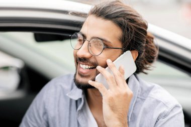 Genç, neşeli, orta doğulu bir sürücünün araba sürerken, yan görünüm, fotokopi çekerken telefon görüşmesi yaptığı yakın plan portresi. Mutlu Arap adam yaz tatilinin tadını çıkarıyor, arabada oturuyor, akıllı telefondan konuşuyor.