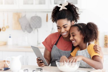 Afro-Amerikan anne ve kızı evde birlikte yemek pişiriyor, çevrimiçi mutfak blogu izliyor, dijital tablet kullanıyor, mutlu siyah anne ve genç çocuk hamur işi yapıyor, ped 'de selfie çekiyor.