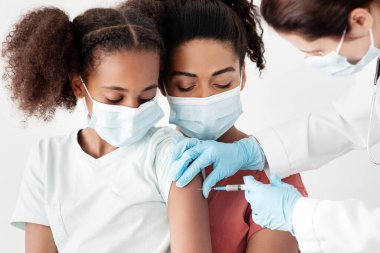 Siyahi bir aile, genç bir anne ve güzel bir kız klinikte aşı yapıyorlar, tıbbi maske takıyorlar. Annesi kucağında oturan kıvırcık saçlı Amerikalı kız için COVID-19 iğnesi yapan hemşire.