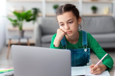 Okul için çok yorgunum. Sıkılmış öğrenci kız çocuğunun portresi masada oturuyor, ödevlerini yapıyor, defterine yazıyor ve dizüstü bilgisayarına bakıyor, online çalışıyor, seçici odaklanıyor. Ev Tabanlı Öğrenme