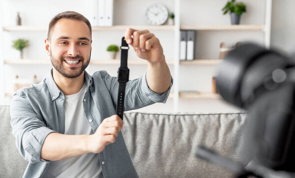 Дружелюбный мужчина-блоггер снимает видео обзор новой модели смарт-часов, используя профессиональную камеру дома. Счастливый контентмейкер, продвигающий современный гаджет, транслирует в соцсети