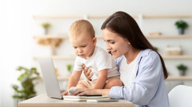 Dizüstü bilgisayarda çalışan ve evde bebeğe bakan gülümseyen genç bir kadın, serbest çalışan bir bayan, bebek oğluyla ve bilgisayarla vakit geçiriyor, doğum izni sırasında uzaktan kumandalı işten hoşlanıyor, boşluğu kopyalıyor.