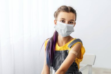 Coronavirus aşısı. Klinikte aşı yaptıktan sonra koluna sargı bezi yapıştırılmış kamera pozu veren koruyucu maskeli ergen öğrenci portresi.
