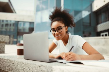 E-öğrenme. Neşeli Afrikalı Öğrenci Laptop 'ta not alıyor. Şehir kolej binasının yakınında uzaktan öğreniyor, gözlük takıyor. Modern Çevrimiçi Eğitim
