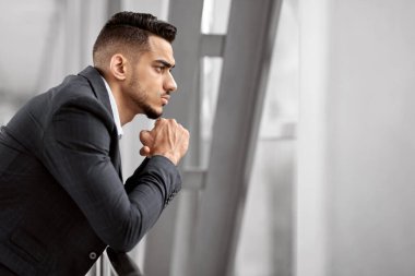 Havaalanında beklerken pencereden bakan düşünceli bir Arap iş adamının portresi parmaklıklara yaslanan ve çenesine dokunan, bir şeyler düşünen, yan görünümü olan dalgın bir Orta Doğulu adam.