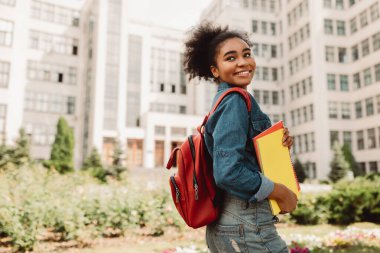 Sırt çantalı, gülümseyen kitaplarla poz veren Afro-Amerikalı Öğrenci Kız Açık Üniversite Binası 'nın yanında duruyor. Modern Eğitim ve Öğrencilik Yaşam Tarzı