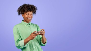 Neşeli Afro-Amerikalı genç adam cep telefonuyla mobil video oyunları oynuyor mavi stüdyonun arka planında duruyor, boş yer var. Duygusal siyahi genç video izliyor, internette geziniyor.